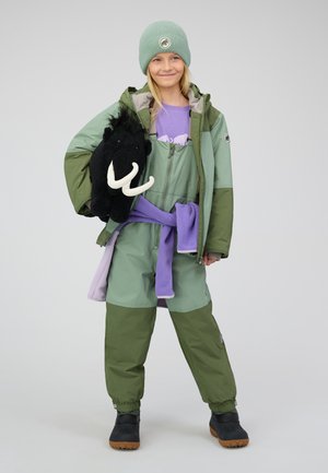 Giacca invernale verde sopra un outfit viola, con in mano un peluche di mammut nero. Include stivali in ecopelle e un berretto verde chiaro.