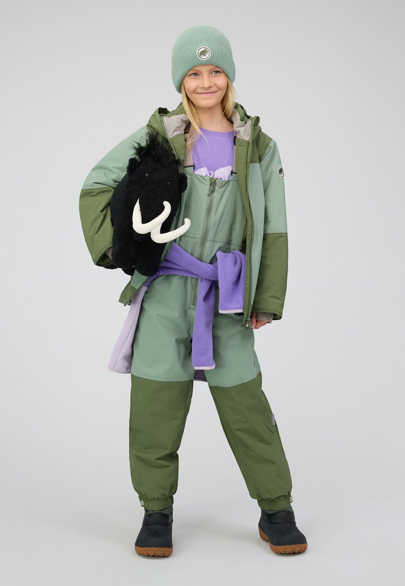 Grüne Winterjacke über einem lila Outfit, hält ein schwarzes Mammut-Plüschtier. Enthält Kunstlederstiefel und eine hellgrüne Mütze.