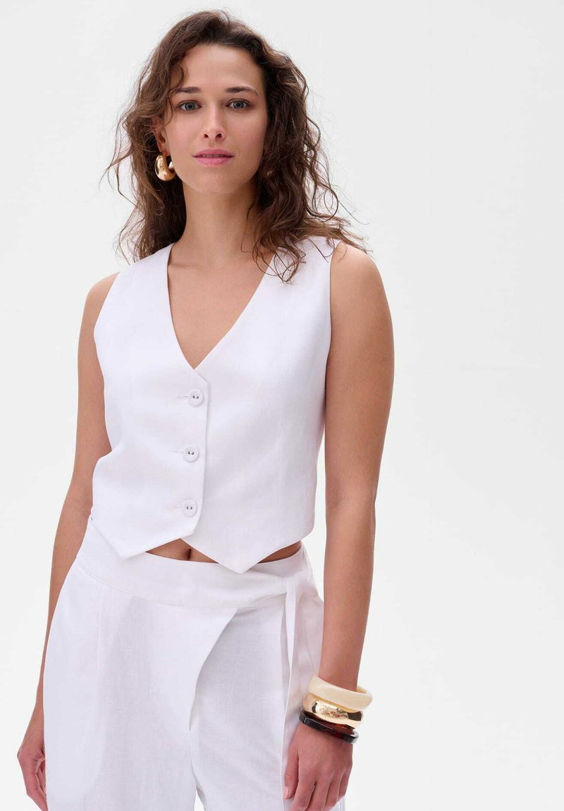 Biały lniany kamizel z głębokim dekoltem w kształcie V, trzema guzikami i dopasowanym krojem. Połączony z luźnymi białymi spodniami i piętrzącymi się bransoletkami na nadgarstku.