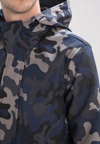 Námořnicky modrá a šedá maskáčová bunda s hladkou texturou, nastavitelnou kapucí a klokáním kapsou. Má zapínání na zip a detailní prošití.