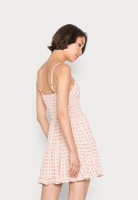 Vestido de verão floral com alças finas, corpete franzido e saias largas. Apresenta um tecido rosa claro com pequenas flores laranja e uma bainha com babados.