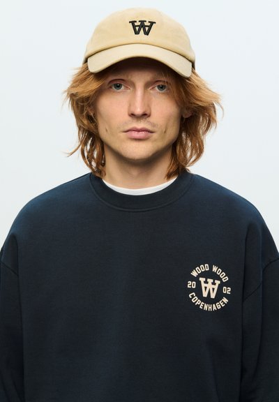 Sweatshirt bleu marine foncé avec un col arrondi, présentant un logo crème sur la poitrine. Associé à une casquette beige arborant un logo "W" noir.