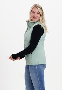 Helles grünes ärmelloses Hemd mit einem gesteppten Design, Reißverschluss, aus glattem Material. Über einem schwarzen Langarmshirt getragen.