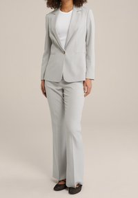 Ljusgrå skräddarsydd blazer och matchande byxor i ett glatt tyg. Blazern har en enda knappstängning och slagkrage. Platta skor synliga.