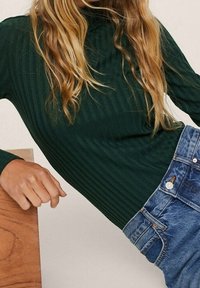 Dunkelgrünes, geripptes Langarmoberteil mit einem Stehkragen, kombiniert mit hellblauen, hoch taillierten Jeans aus Denim mit Fronttaschen und einem Metallknopf.