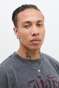 Et nærbilde av en person med kort hår som har på seg en falmet svart grafisk t-skjorte og en sølvkjede med et anheng. Merkbare tatoveringer er synlige.
