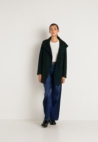 Mulher vestindo um casaco verde-escuro sobre uma t-shirt branca cropped, jeans de perna larga azuis e sapatos pretos, de pé contra um fundo branco liso.