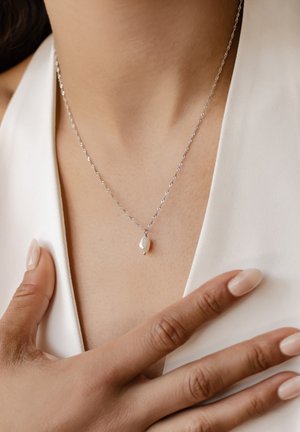 Collana in argento a catena intrecciata con un ciondolo in perla bianca ovale, posizionata sopra la clavicola di una persona, con una leggera texture della pelle visibile.