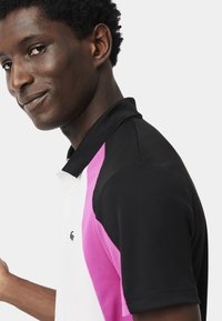 Polo noir et blanc avec des accents roses, doté d'un col classique et de manches courtes. Comprend un petit logo vert sur la poitrine.