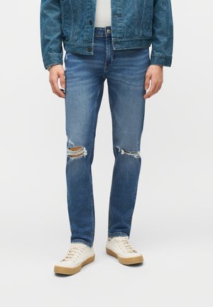 Homme portant un jean bleu délavé moyen déchiré, des baskets blanches à semelles en caoutchouc et une veste en denim bleue à motifs, debout devant un fond uni.