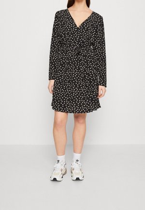 Femme portant une robe noire à manches longues avec un petit imprimé floral blanc, des chaussettes blanches et des baskets blanches, debout devant un fond uni.