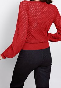 Czerwona, teksturowana sweter z diamentowym wzorem, długimi rękawami i ściągaczem na dole. Noszony z ciemnoniebieskimi dżinsami, ukazując dopasowaną sylwetkę.