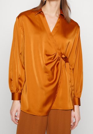 Femme portant un chemisier cache-cœur en satin orange brûlé brillant, avec de longues manches bouffantes et un détail noué à la taille.