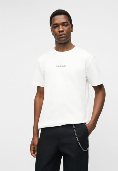 Νέος άνδρας που φορά λευκό κοντομάνικο T-shirt με το κείμενο "The Kooples" και μαύρο παντελόνι με αξεσουάρ ασημένιας αλυσίδας.