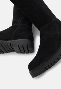 Stivali alti fino al ginocchio in suede nero con ampia suola a piattaforma, dotati di zip laterale e suola in gomma testurizzata per una migliore aderenza.