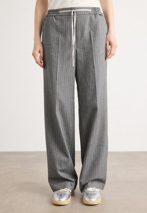 Personne portant un pantalon gris à fines rayures ample avec une taille à cordon et des baskets argentées, debout sur un sol gris.