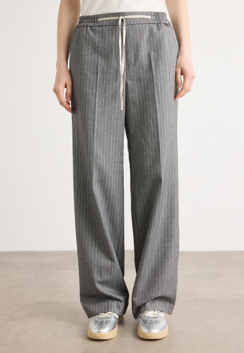 Personne portant un pantalon gris à fines rayures ample avec une taille à cordon et des baskets argentées, debout sur un sol gris.