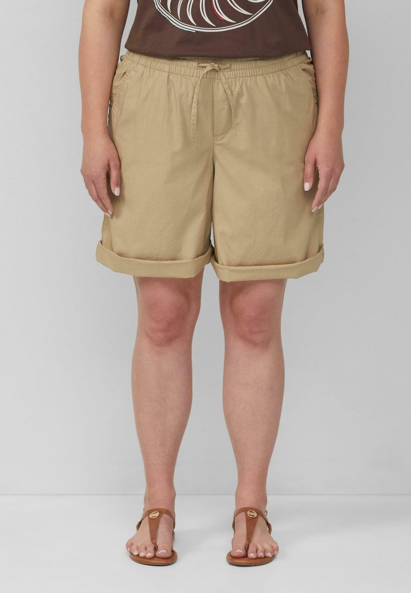 Beige Baumwollshorts mit elastischem Bund und Kordelzug, mit umgeschlagenen Säumen und Seitentaschen. Getragen mit braunen Sandalen.