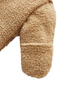Tan farbener Plüschbär mit einem Arm aus weichem, strukturiertem Stoff mit einem flauschigen Finish; abgerundete Spitze und sichtbare Nähte.
