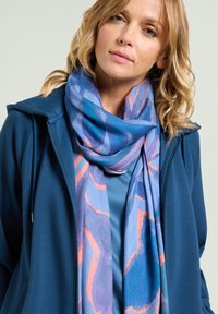 Sudadera con capucha azul con cremallera, con una camiseta azul de capas debajo, combinada con una bufanda ligera con un estampado floral en tonos azules y naranjas.