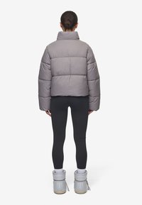 Kvinde med hår i en knold iført en grå pufferjakke, sorte leggings, hvide sokker og lysegrå vinterstøvler, stående med ryggen til på hvid baggrund.