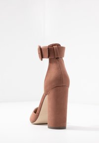 Brown suede high-heeled shoe with a chunky heel and ankle strap, featuring a square buckle and smooth texture.