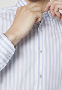 Jasnoniebiesko-biała koszula w paski wykonana z tkaniny o gładkiej fakturze; posiada klasyczny kołnierzyk oraz guziki z przodu i na mankietach.