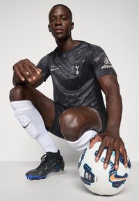 T-shirt de sport noir à motifs avec manches courtes, logo Nike, et shorts blancs. Le joueur tient un ballon de soccer multicolore, portant des crampons noirs.