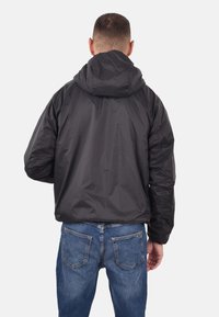 Giacca nera leggera con cappuccio, dal tessuto liscio e con polsini elasticizzati, indossata sopra jeans di denim blu, vista da dietro.