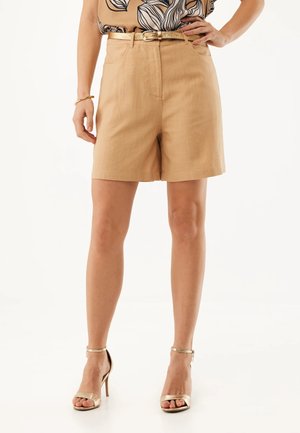 Kobieta w beżowych szortach z wysokim stanem i złotym paskiem, w połączeniu z wzorzystą bluzką i złotymi sandałami na wysokim obcasie, stojąca z ręką na biodrze.