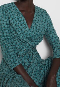 Robe portefeuille bleu sarcelle à manches trois-quarts, présentant un motif tourbillonnant dans des nuances plus foncées et une ceinture nouée à la taille pour une silhouette ajustée.