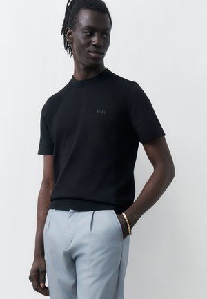 Άνδρας που φοράει μαύρο κοντομάνικο μπλουζάκι BOSS και ανοιχτό γκρι παντελόνι, με το ένα χέρι στην τσέπη και ένα χρυσό βραχιόλι στον καρπό.