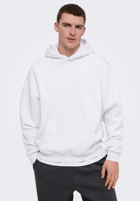 Jeune homme portant un sweat à capuche blanc uni et un pantalon foncé, debout avec une main dans la poche sur un fond clair.