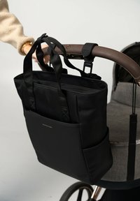 Borsa tote in tessuto nero con manici doppi, dotata di una tasca frontale e attaccata al manico in pelle marrone di un passeggino. Texture liscia, design strutturato.