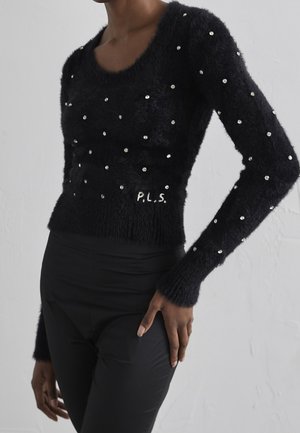 Pull noir duveteux à manches longues avec de petites décorations argentées et une broderie "P.L.S.", porté avec un pantalon noir ajusté.