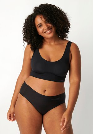 Donna sorridente con capelli ricci che indossa un reggiseno nero senza cuciture e biancheria intima abbinata su uno sfondo chiaro.