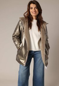 Veste matelassée argent métallique avec doublure en fausse fourrure, fermeture éclair à l'avant et capuche. Associée à un léger pull et un jean large.
