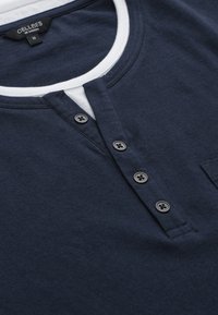 Mörkblå bomulls-Henley-tröja med vit rund halsringning, fem knappar framtill och en bröstficka för extra detalj. Mjuk textur.