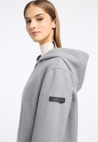 Grijze hoodie met een zachte textuur, voorzien van een rits en een zwart label op de mouw, gecombineerd met een geribbelde beige coltrui.