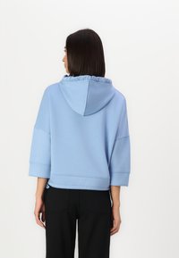 Felpa blu corta con cappuccio arricciato e maniche ampie, realizzata in tessuto morbido. Abbinata a pantaloni neri con dettagli sulle tasche posteriori.