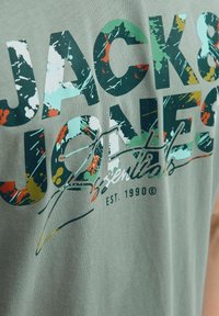 Grøn bomulds t-shirt med dristig, multicolor tekst og sprøjtede mønster. Teksten inkluderer "JACK & JONES" og "essentials" med et signaturdesign.