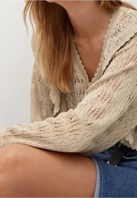 Femme portant un haut en tricot beige à manches longues avec des bords festonnés et un short en jean bleu, assise avec un bras reposant sur son genou.