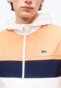 Νεαρός που φοράει αθλητικό μπουφάν με φερμουάρ, με πορτοκαλί, ναυτικό μπλε και λευκά χρώματα και πράσινο λογότυπο κροκόδειλου στο στήθος.