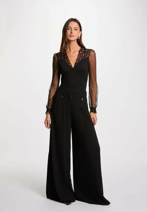 Body nero con maniche lunghe trasparenti a pois e rifiniture in pizzo, abbinato a pantaloni neri a gamba larga con pieghe e hardware minimal.