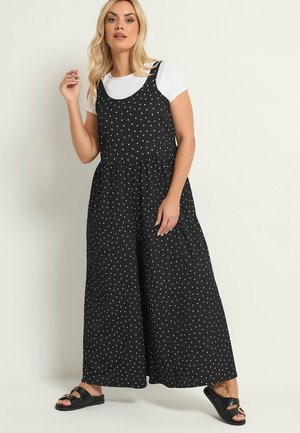 Lyshåret kvinde iført en sort ærmeløs jumpsuit med hvide prikker over en hvid t-shirt og sorte slip-on sandaler, stående mod en ensfarvet baggrund.