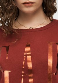 Κοντινό πλάνο ενός ατόμου που φοράει κόκκινο πουκάμισο με μεταλλικά γράμματα και χρυσό κολιέ με μικρό μενταγιόν.