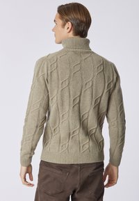 Maglione beige a trecce con colletto alto. Presenta pattern testurizzati e polsini e orlo a coste. Indossato con pantaloni marroni.