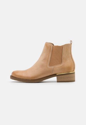 Enkelhoge laars van beige leer met elastisch zijpaneel, lage gestapelde hak en gouden accent boven de hiel op een witte achtergrond.