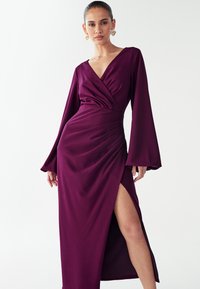 Lila langermige jurk met een diepe V-hals, geplooide taille en een hoge split, gemaakt van een soepele, vloeiende stof.