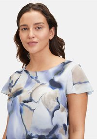 Fließende Bluse in Hellblau mit abstrakten Aquarellmustern. Mit runder Halsaussschnitt und kurzen Rüschensäumen. Weicher Stoff.